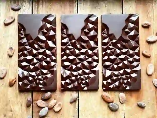 Three Neary Nógs chocolate bars with geometric textures, surrounded by cacao beans on a rustic wooden table.
