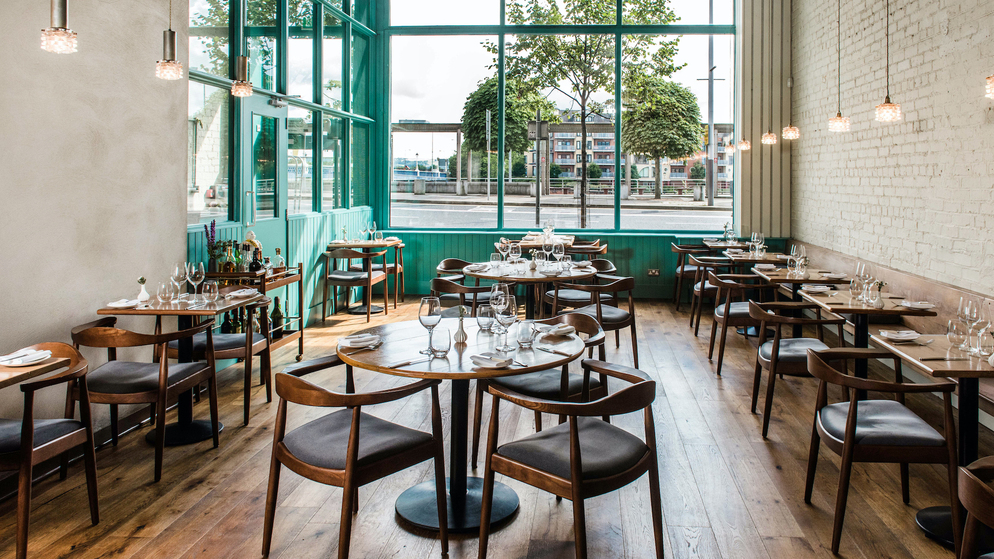 Dining room at OX Belfast with wooden tables, teal-framed windows and views towards the River Lagan.