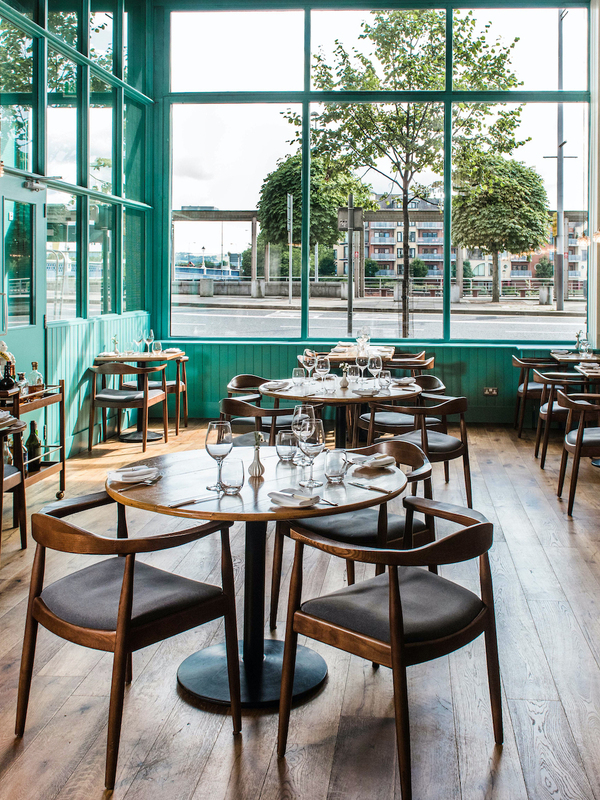 Dining room at OX Belfast with wooden tables, teal-framed windows and views towards the River Lagan.