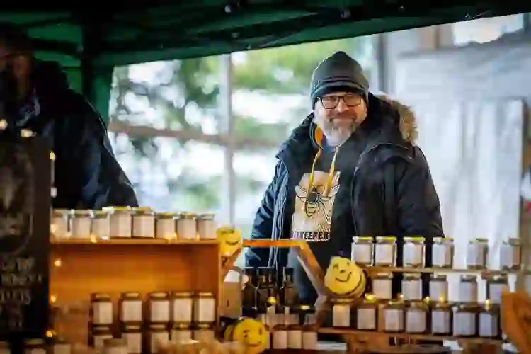 A beekeeper wearing a woolly hat stands behind a stall at Mahon Point Farmers' Market, County Cork, displaying rows of jars of honey.