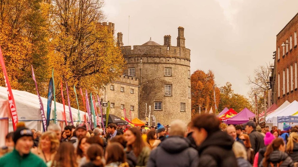 Menschenmengen stöbern an bunten Essensständen vor der Burg von Kilkenny während des Savour Kilkenny Food Festival im Herbst.