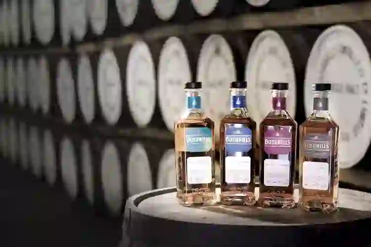 Bottles of Bushmills Irish whiskey displayed on a barrel in front of rows of ageing whiskey casks.