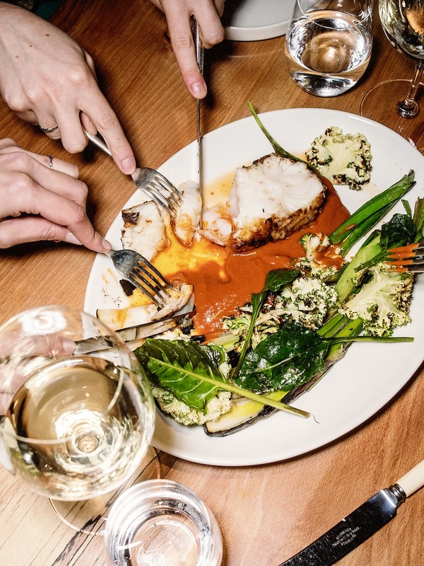 Diners share a seafood dish with grilled vegetables and wine at a wooden table.