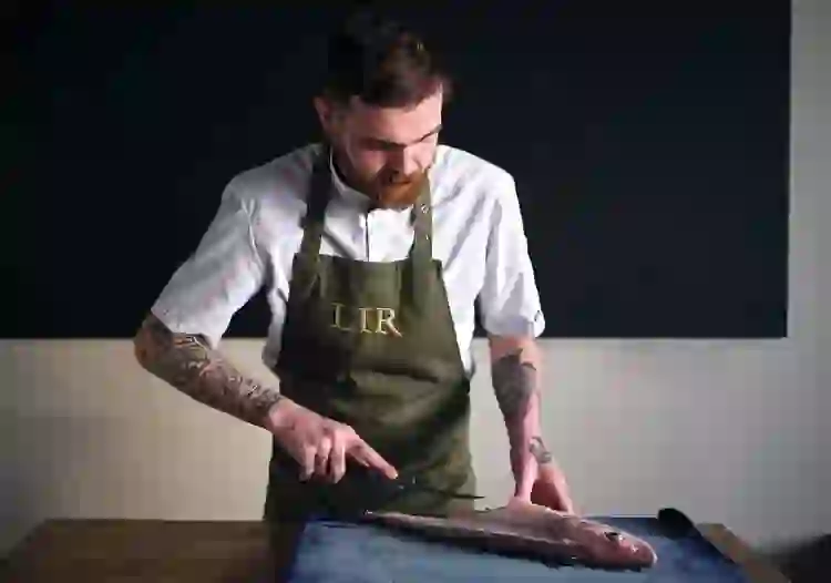 Ein Koch in einer weißen Jacke und Lir-Schürze filetiert einen ganzen Fisch auf einem Schneidebrett.