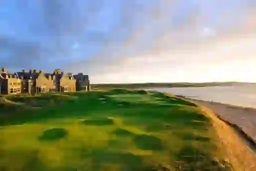 The coastal fairway at Doonbeg, County Clare, with a grand stone lodge beside the sea, lit by warm evening sunlight.