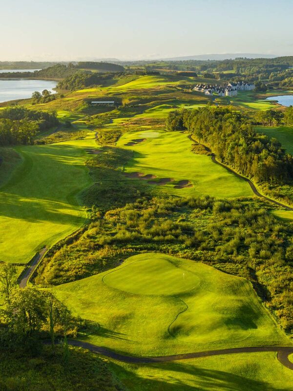 Vue aérienne panoramique du Lough Erne Golf Resort avec des fairways verts, des lacs et des collines environnantes au coucher du soleil.