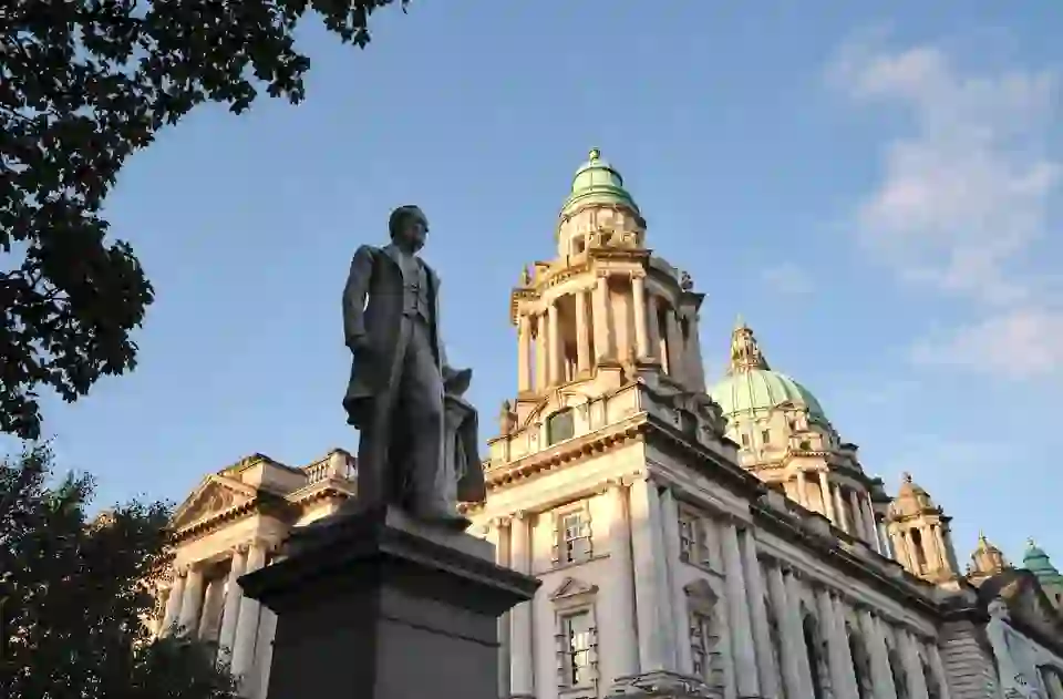 Eine Statue steht vor den prächtigen Kuppeln und der kunstvollen Fassade des Rathauses von Belfast unter einem klaren Abendhimmel.