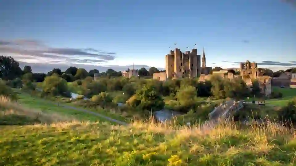 Trim Castle rises above the River Boyne at sunset, surrounded by green parkland and historic ruins in County Meath.