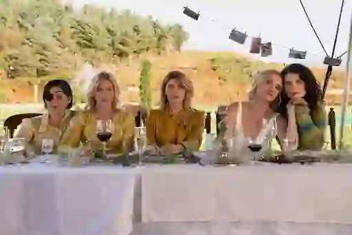 Five women from Bad Sisters sit at a long outdoor table with wine glasses, staring ahead in silence.