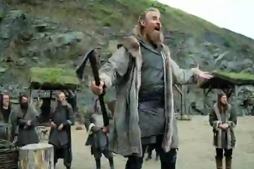 Bearded Viking leader holds an axe and addresses a crowd in a rocky outdoor camp in Vikings: Valhalla.