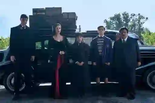 The Addams family stand in front of a black car with luggage stacked on the roof in Wednesday.
