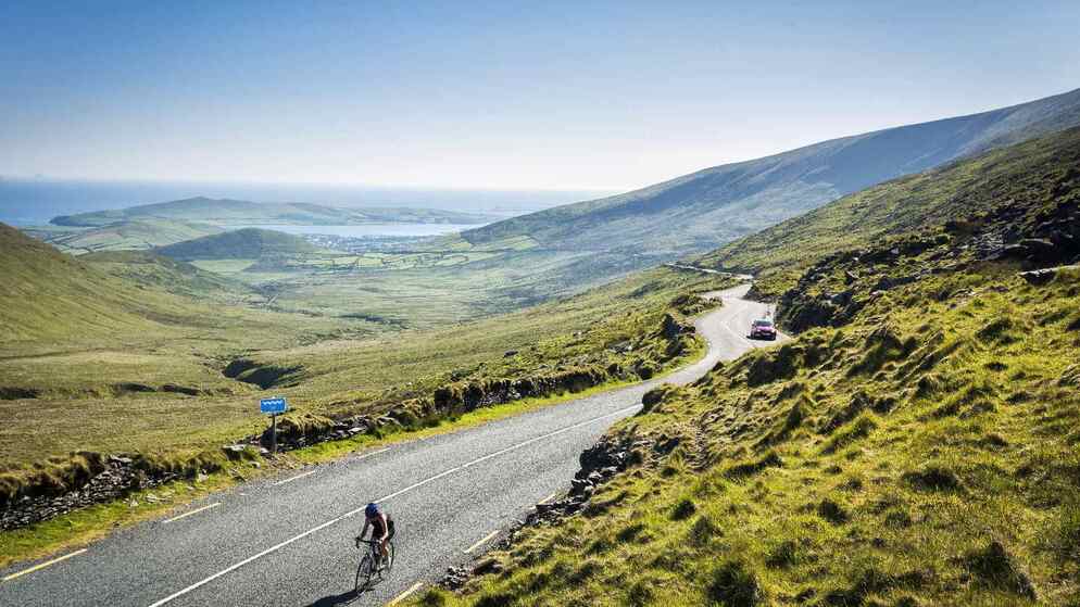 A winding coastal road curves through green hills overlooking Dingle Bay, with sweeping ocean views under a clear blue sky.