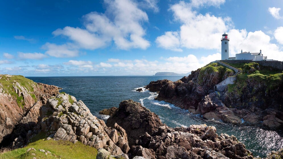 Fanad Head Lighthouse perched on a rocky cliff overlooking the Atlantic Ocean under a bright blue sky with scattered clouds.