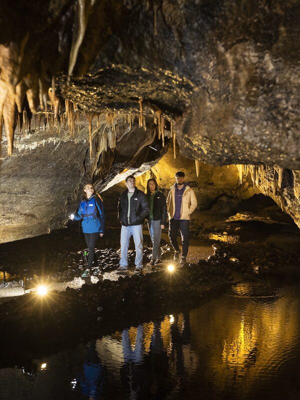 Group exploring a limestone cave with stalactites in Ireland, lit by warm lights reflecting on underground pools.