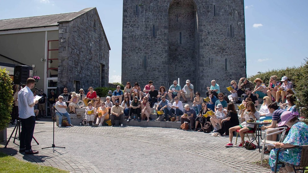 Crowd reading on steps outside medieval stone building during a sunny event at an Irish literary festival.