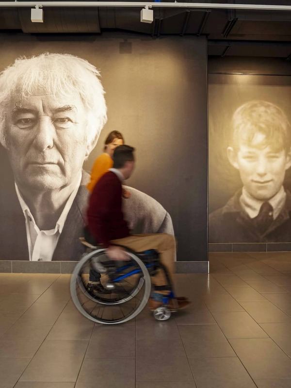 Portraits of Seamus Heaney and his younger self at HomePlace; visitors pass by, immersed in his story.