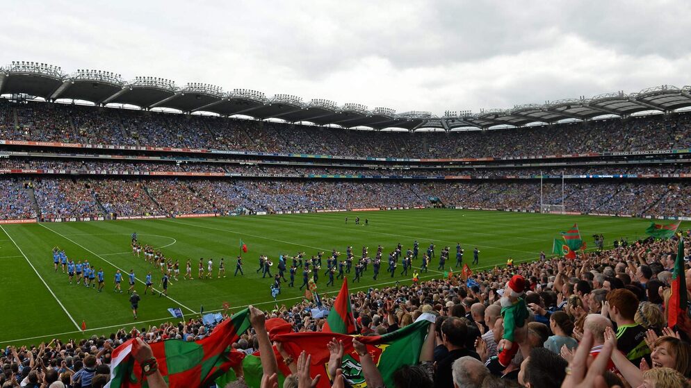 Full crowd at Croke Park in Dublin watching pre-match parade before a Gaelic football final, with fans waving county flags.