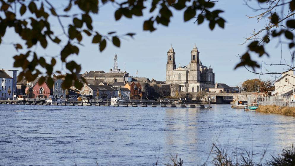 Vista sul fiume Shannon verso le casette a schiera di Athlone e le due guglie della chiesa, incorniciate da rami frondosi.