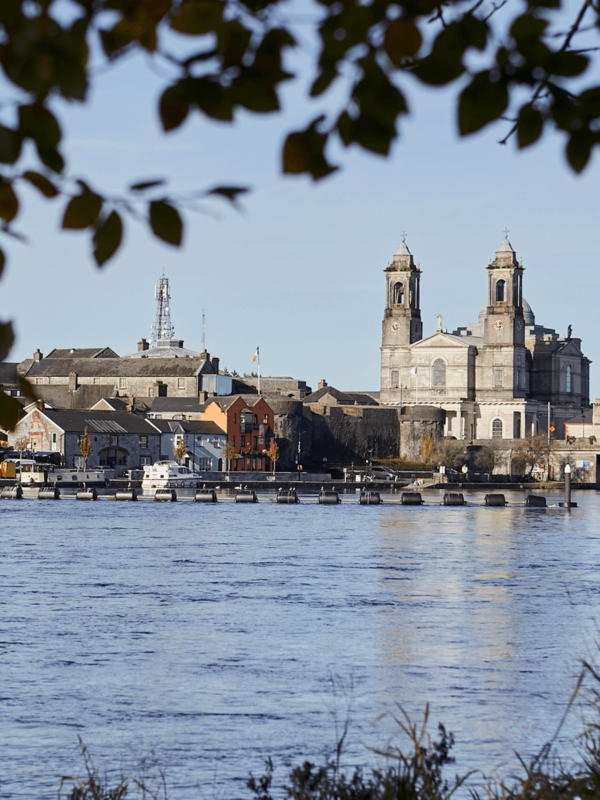 Uitzicht over de rivier Shannon op de rijtjeshuizen van Athlone en de twee kerktorens, omlijst door bladerrijke takken.