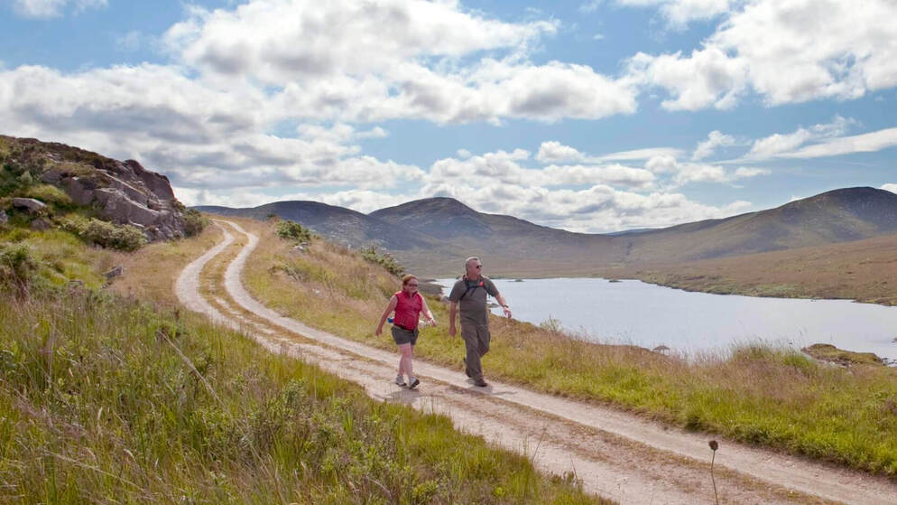 Two walkers follow a winding trail along Lough Veagh in County Donegal, with rugged hills under a cloud-filled summer sky.