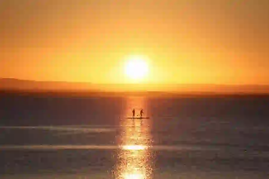 Two figures paddleboarding across shimmering water at sunset.