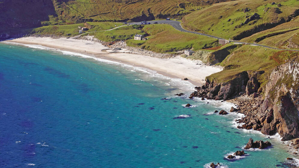 Black-faced sheep graze on a grassy slope above the turquoise waters and sandy shore of Keem Bay on Achill Island.