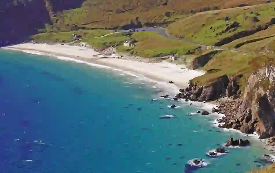 A curving stretch of white sand and turquoise water lies beneath rugged cliffs and grassy hills.