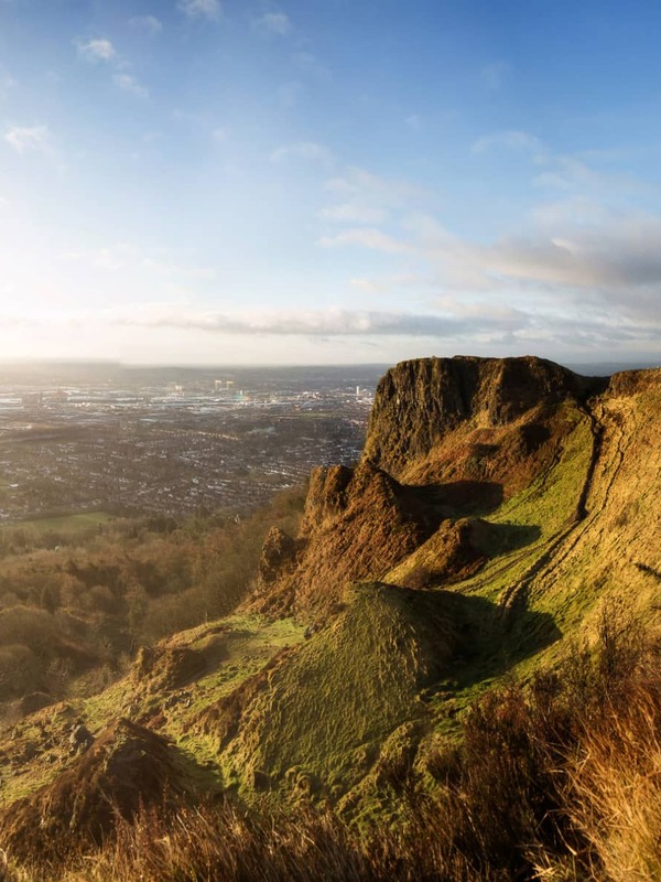Wide view of Cave Hill’s rugged cliffs at sunset overlooking Belfast city and Lough in the distance.
