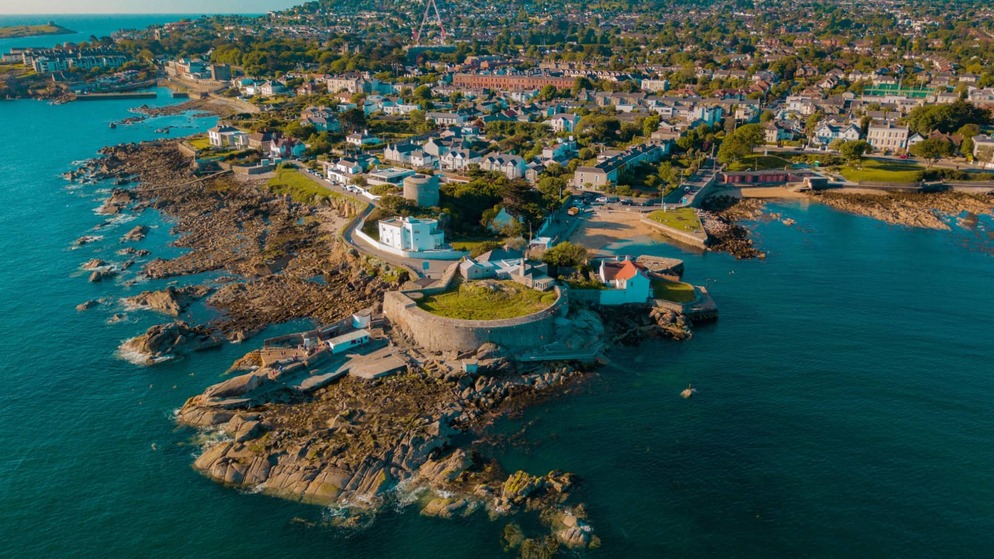 Aerial view of coastal houses and rocky shoreline at the Forty Foot swimming spot near Dublin, with blue sea and harbour.