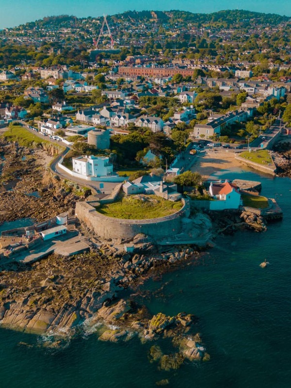 Aerial view of coastal houses and rocky shoreline at the Forty Foot swimming spot near Dublin, with blue sea and harbour.