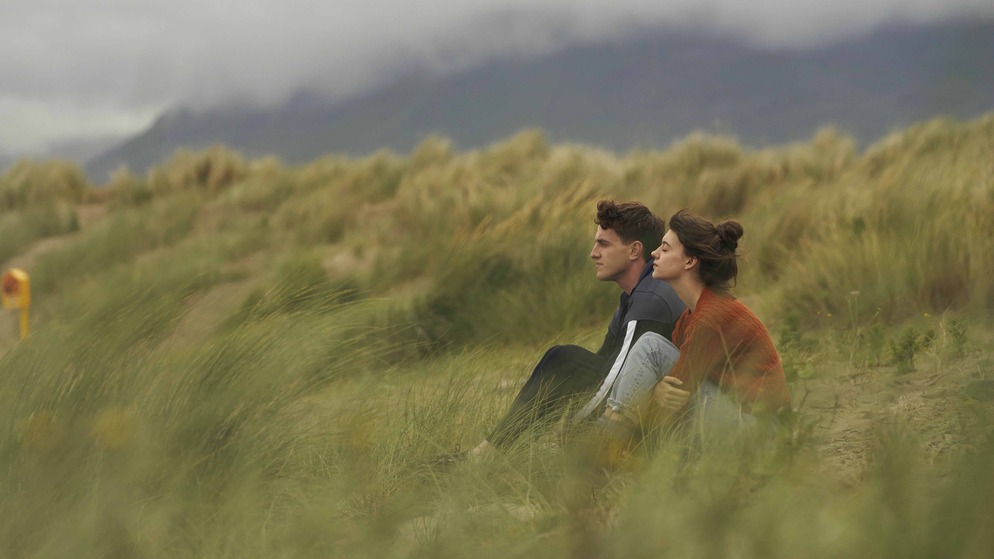 Marianne and Connell sit in sand dunes with misty mountains in the distance in County Sligo.