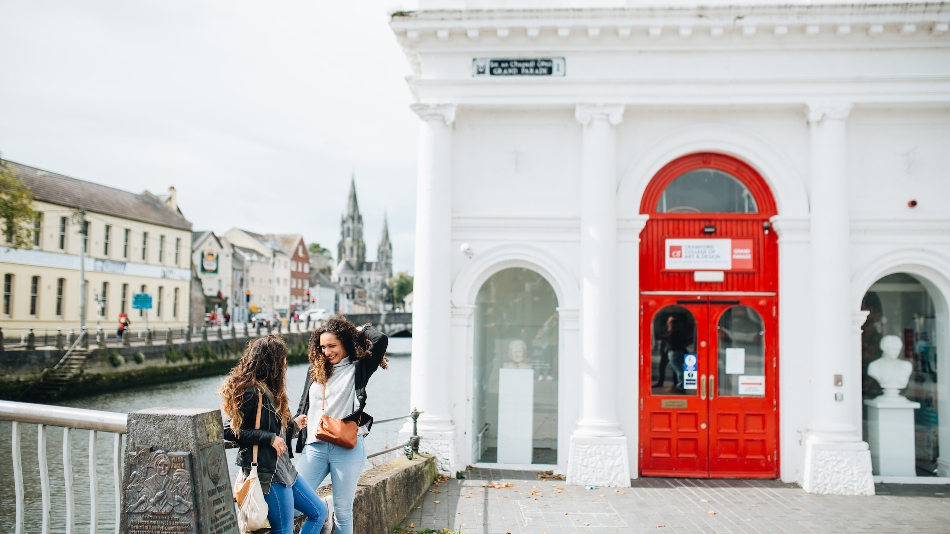 Two women chatting by the River Lee beside the red entrance of the Cork City Gaol museum, with St Fin Barre’s Cathedral in the background.