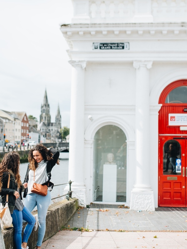 Two women chatting by the River Lee beside the red entrance of the Cork City Gaol museum, with St Fin Barre’s Cathedral in the background.