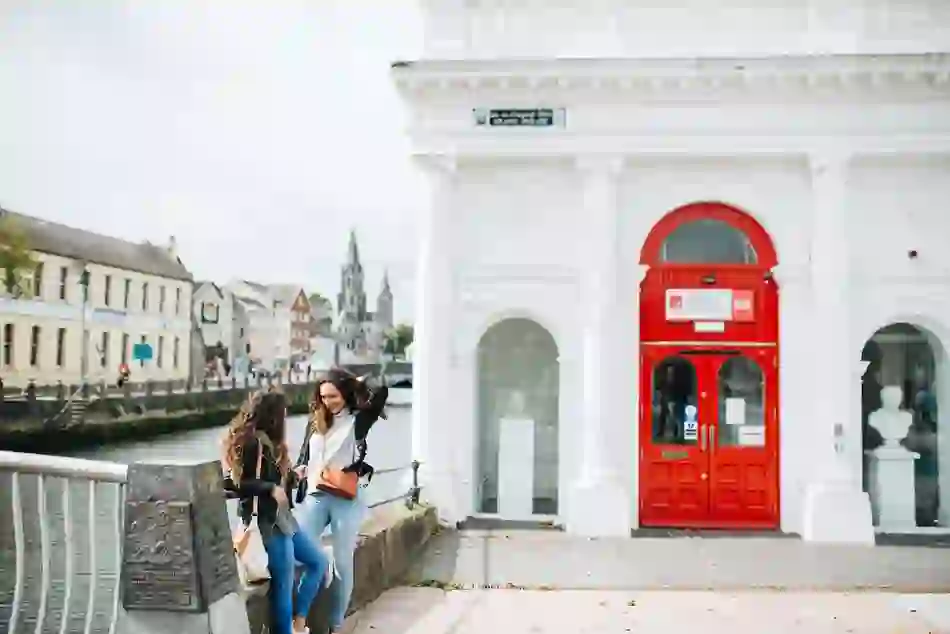 Two women chatting by the River Lee beside the red entrance of the Cork City Gaol museum, with St Fin Barre’s Cathedral in the background.