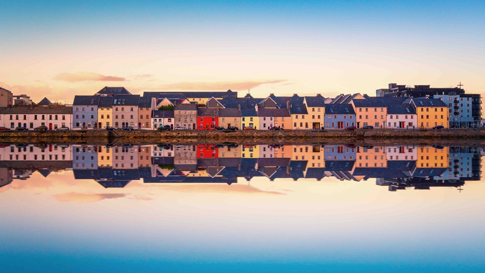 Reihe von pastellfarbenen Häusern, die sich im Wasser am Long Walk in Galway bei Sonnenuntergang spiegeln.