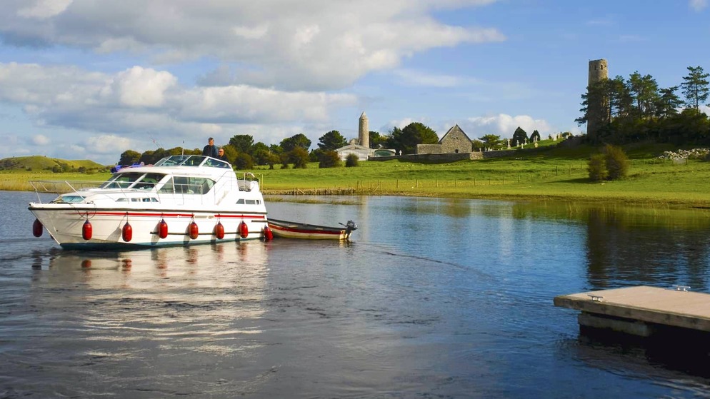 Weißer Motorbootkreuzer auf dem Fluss Shannon mit Clonmacnoise monastery und Rundturm im Hintergrund.