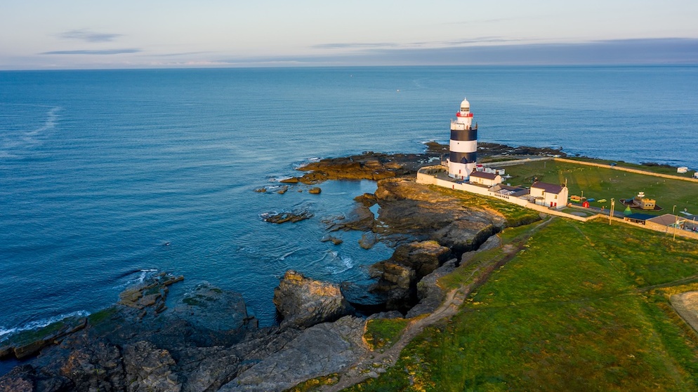 Hook Lighthouse overlooking the sea on the Hook Peninsula, surrounded by outbuildings and rocky shoreline at golden hour.