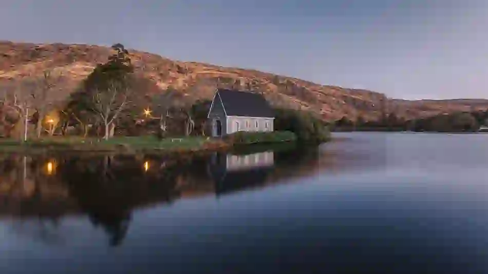 Gougane Barra church beside a calm lake at dusk, with soft reflections and rugged hills in the background.
