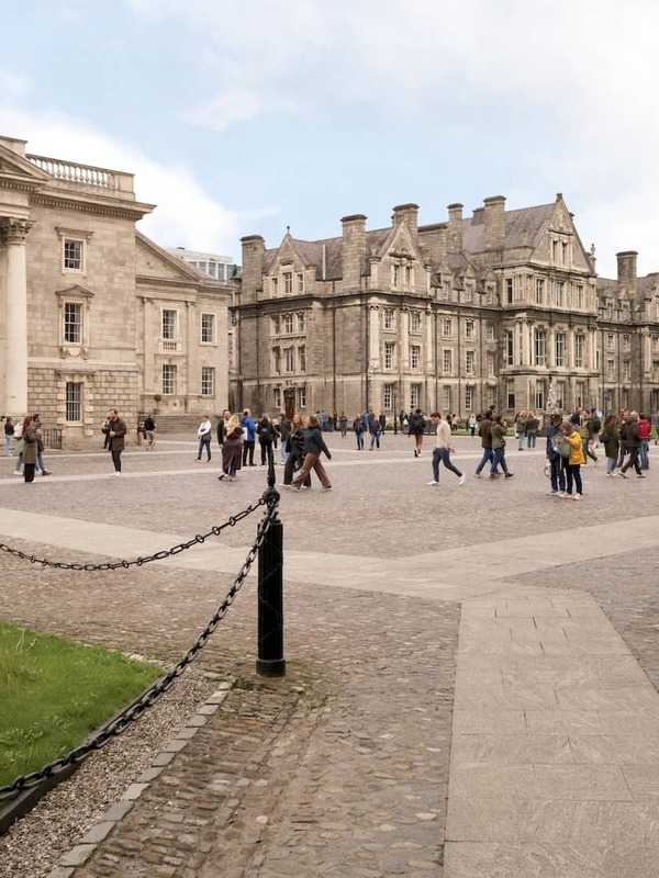 Students and visitors walking through the cobbled front square at Trinity College Dublin with the Campanile and historic buildings.