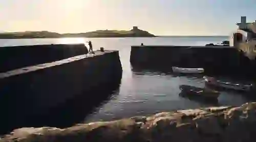 Der Hafen von Colliemore in Dalkey bei Sonnenuntergang: Kleine Boote ruhen im stillen Wasser, und eine Person spaziert entlang der Landungsbrücke.