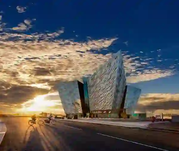 Titanic Belfast bei Sonnenuntergang: Radfahrer fahren vorbei und goldenes Licht liegt über dem Hafen von Belfast.