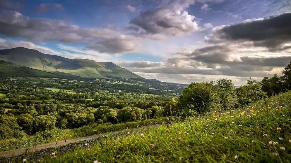 Scenic view of rolling green hills and dramatic mountains under a cloudy sky in Ireland’s Hidden Heartlands.