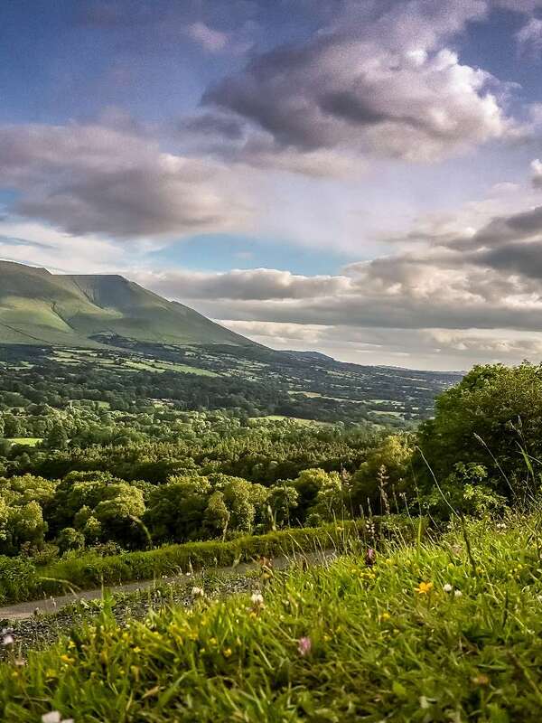 Scenic view of rolling green hills and dramatic mountains under a cloudy sky in Ireland’s Hidden Heartlands.