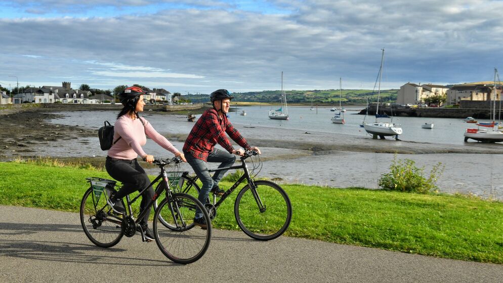 Couple cycling along the Waterford Greenway with boats docked in a calm harbour behind them.