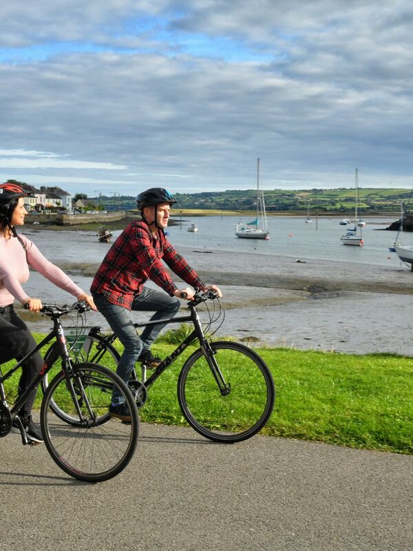 Una coppia in bicicletta lungo il Waterford Greenway con barche ormeggiate in un porto tranquillo dietro di loro.