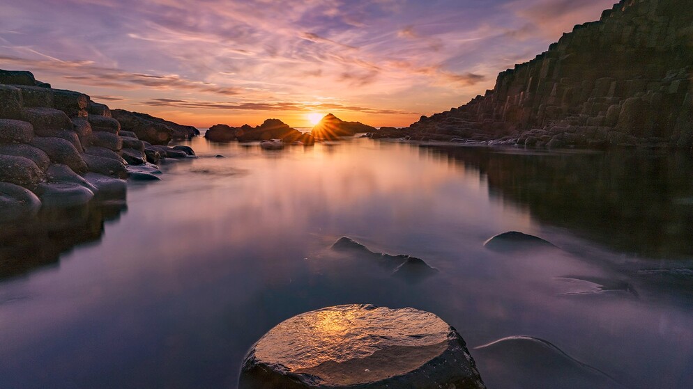Puesta de sol en la Calzada del Gigante con un mar en calma, un cielo resplandeciente y formaciones de basalto negro reflejadas en el agua tranquila.