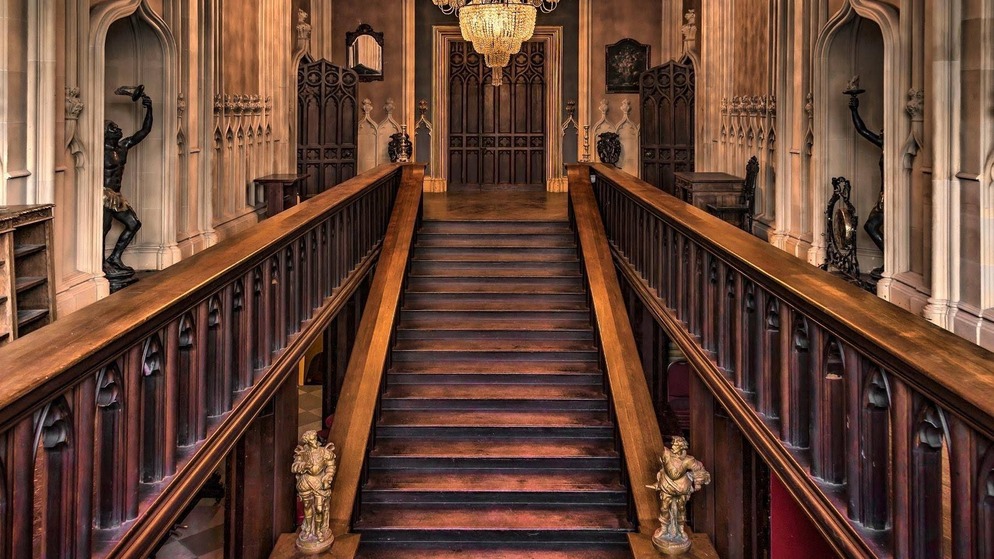 Verzierte Holztreppe mit geschnitzten Paneelen und einem Kronleuchter in einem prächtigen historischen Gebäude.