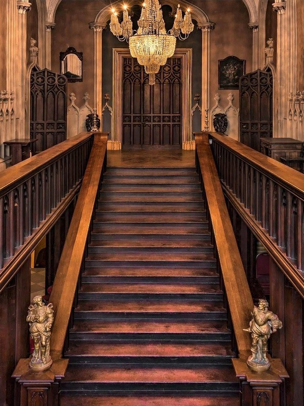 Ornate wooden staircase with carved panels and chandelier inside a grand historic building.