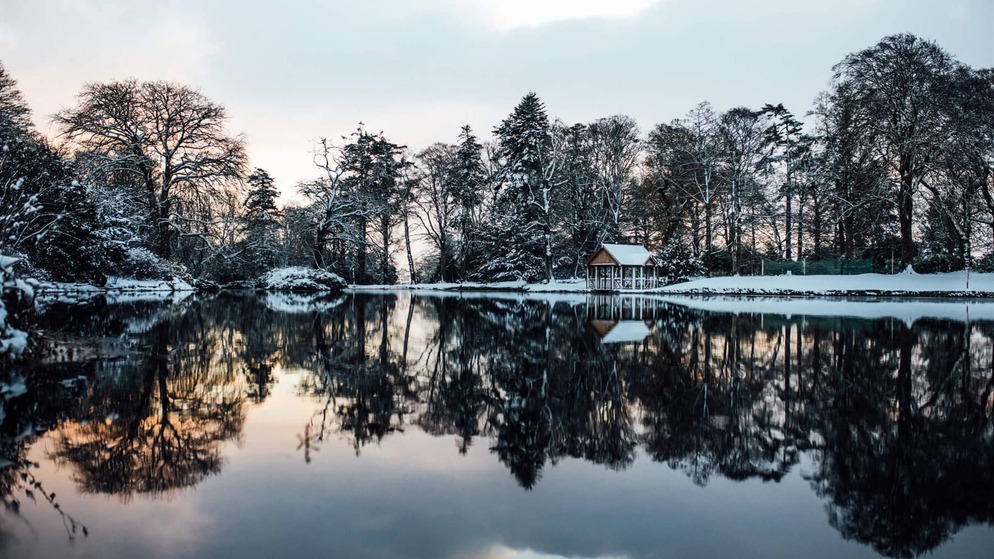 A snowy lakeside hut is mirrored in still waters beneath frost-draped trees at dusk.