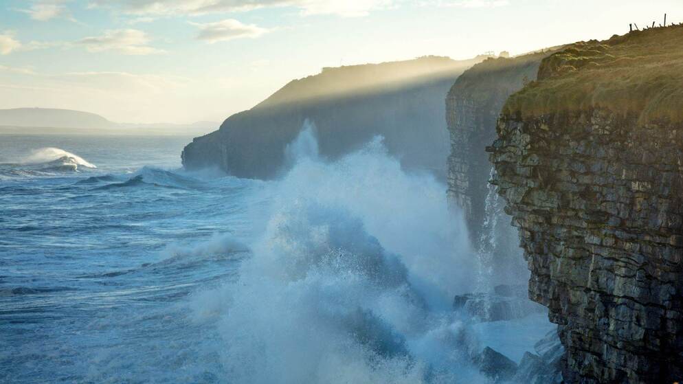 Dramatische Atlantische golven beuken tegen hoge zeekliffen in County Sligo tijdens het gouden uur, terwijl het zonlicht door de mist breekt.